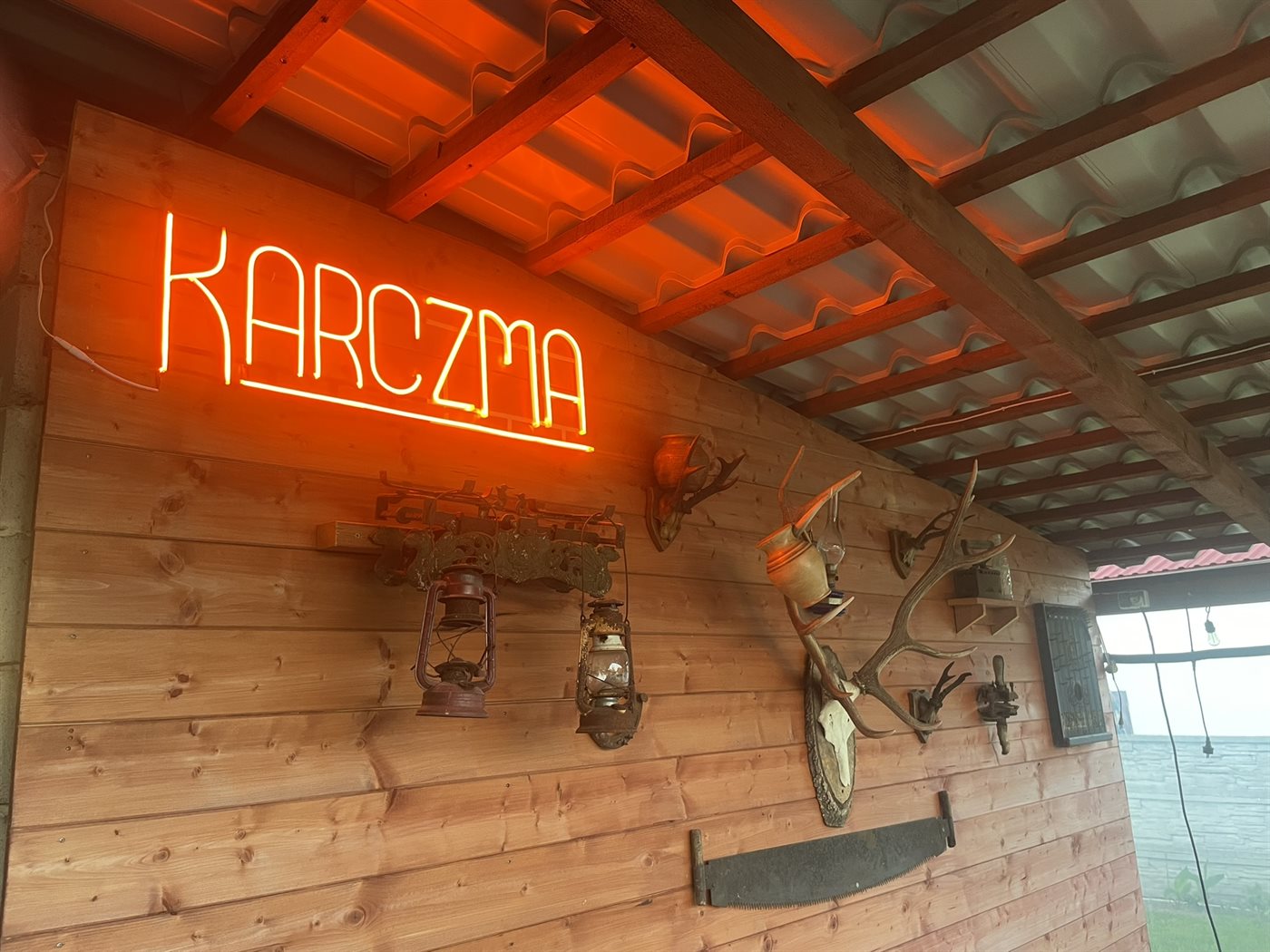 Neon z napisem KARCZMA w drewnianej altanie ogrodowej, ciepłe pomarańczowe światło